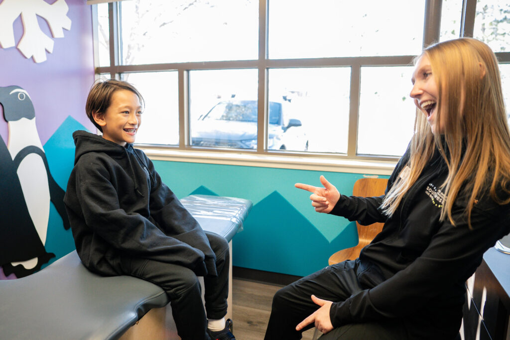 Team member at Southwest Pediatric Dentistry & Orthodontics laughing with a young patient on an exam chair in a kid-friendly room with a penguin mural. The room was designed to help children feel at ease at their Littleton, CO dental visits.